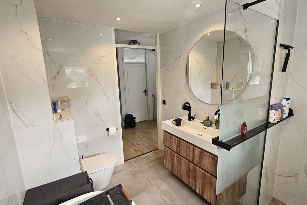 Opgeleverde badkamer met wanden in marmerlook, een ronde spiegel, houten wastafelmeubel en glazen douchewand
